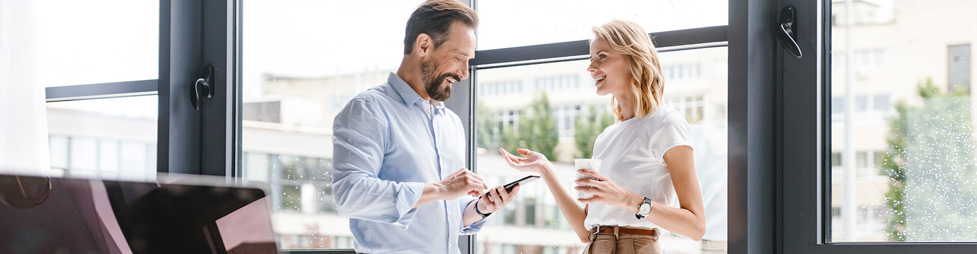 Carteras Premium - Hombre con camisa y móvil en la mano, y mujer de negocios con cafe en la mano sonriendo en la oficina Carteras Premium - Hombre con camisa y móvil en la mano, y mujer de negocios con cafe en la mano sonriendo en la oficina
