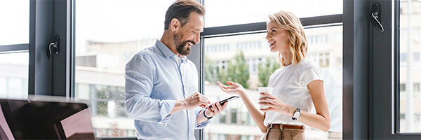 Carteras Premium - Hombre con camisa y móvil en la mano, y mujer de negocios con cafe en la mano sonriendo en la oficina Carteras Premium - Hombre con camisa y móvil en la mano, y mujer de negocios con cafe en la mano sonriendo en la oficina
