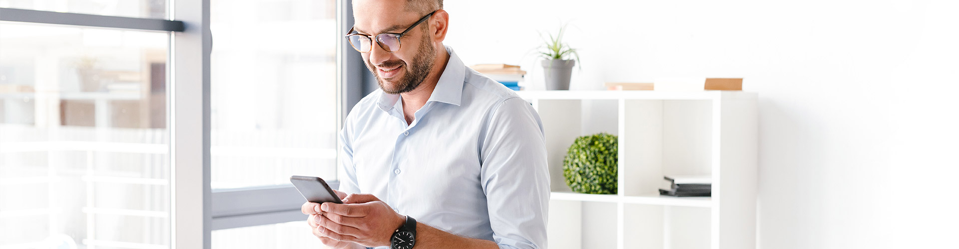 Cuenta a la Vista - Hombre de negocios con camisa y gafas sentado sobre su mesa en la oficina con un movil en la mano sonriendo Cuenta a la Vista - Hombre de negocios con camisa y gafas sentado sobre su mesa en la oficina con un movil en la mano sonriendo