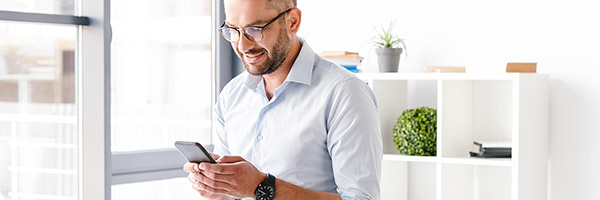Cuenta a la Vista - Hombre de negocios con camisa y gafas sentado sobre su mesa en la oficina con un movil en la mano sonriendo Cuenta a la Vista - Hombre de negocios con camisa y gafas sentado sobre su mesa en la oficina con un movil en la mano sonriendo