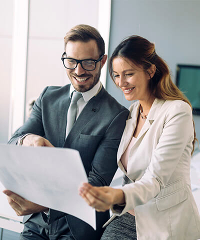 Momento Hacer crecer mi empresa - Hombre y mujer de negocios con traje, corbata y gafas sonriendo mientras miran papeles en la oficina Momento Hacer crecer mi empresa - Hombre y mujer de negocios con traje, corbata y gafas sonriendo mientras miran papeles en la oficina