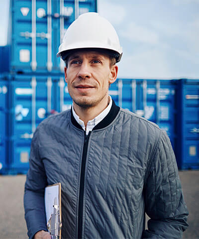 Momento Comenzar a exportar e importar - Trabajador con casco blanco y chaqueta azul sonriendo mientras trabaja en un puerto rodeado de contenedores Momento Comenzar a exportar e importar - Trabajador con casco blanco y chaqueta azul sonriendo mientras trabaja en un puerto rodeado de contenedores