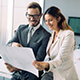 Momento Hacer crecer mi empresa - Hombre y mujer de negocios con traje, corbata y gafas sonriendo mientras miran papeles en la oficina Momento Hacer crecer mi empresa - Hombre y mujer de negocios con traje, corbata y gafas sonriendo mientras miran papeles en la oficina