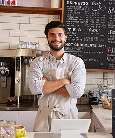 Ventajas Ampliar o reformar mi negocio - Dueño joven de una cafeteria con delantal y los brazos cruzados sonriendo tras la barra Ventajas Ampliar o reformar mi negocio - Dueño joven de una cafeteria con delantal y los brazos cruzados sonriendo tras la barra