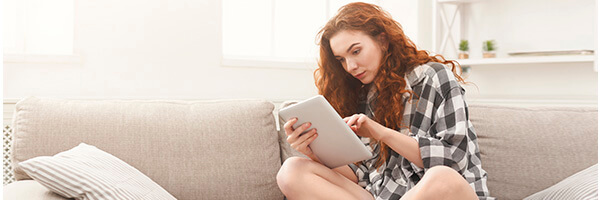 Cuenta de Pago Básica - Chica joven con una camisa de cuadros sentada en el sofá de su casa con una tablet buscando cuentas Cuenta de Pago Básica - Chica joven con una camisa de cuadros sentada en el sofá de su casa con una tablet buscando cuentas