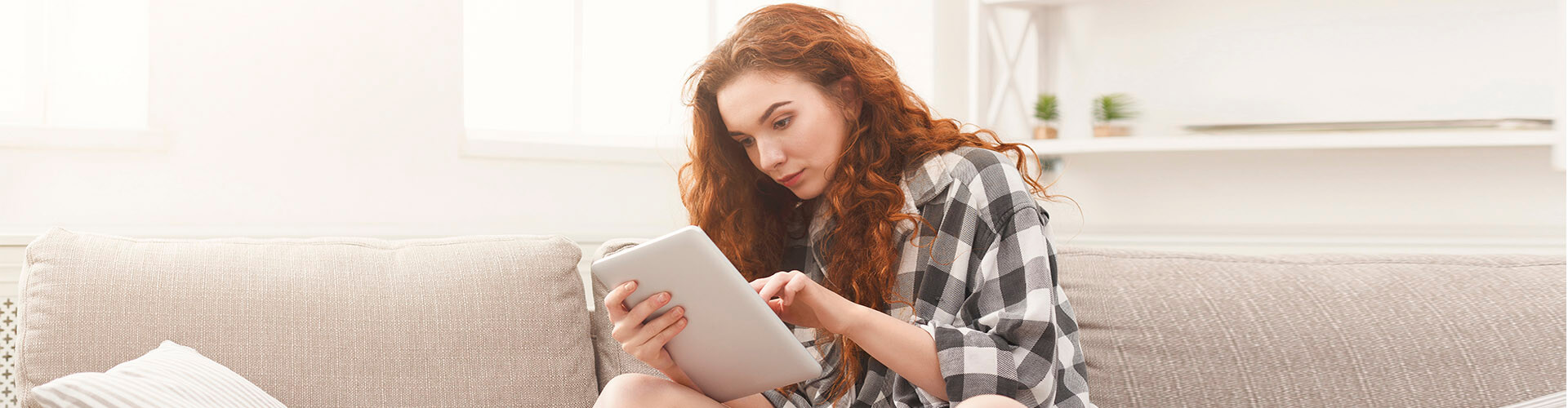 Cuenta de Pago Básica - Chica joven con una camisa de cuadros sentada en el sofá de su casa con una tablet buscando cuentas Cuenta de Pago Básica - Chica joven con una camisa de cuadros sentada en el sofá de su casa con una tablet buscando cuentas