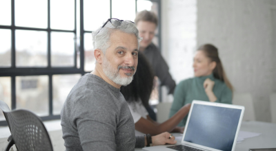 Señor sentado en oficina mirando a cámara con dos compañeros desenfocados al fondo