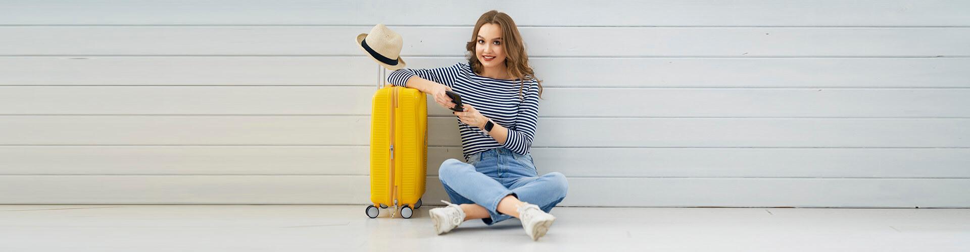 Seguro de Asistencia en Viaje - Mujer joven sentada en el suelo con el movil en la mano, junto a su maleta y un sombrero preparando un viaje