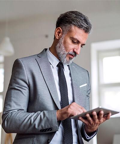 Momento Digitalizar mis gestiones- Hombre de mediana edad con barba, traje gris y corbata negra trabajado en la oficina con una tablet pensando en digitalizar las gestiones de su empresa