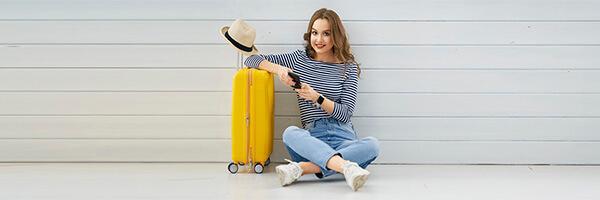 Seguro de Asistencia en Viaje - Mujer joven sentada en el suelo con el movil en la mano, junto a su maleta y un sombrero preparando un viaje Seguro de Asistencia en Viaje - Mujer joven sentada en el suelo con el movil en la mano, junto a su maleta y un sombrero preparando un viaje