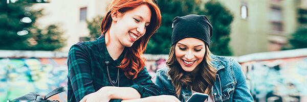 Programa Joven In - Dos chicas jovenes sonriendo mientras navegan por intenet con el movil en un parque Programa Joven In - Dos chicas jovenes sonriendo mientras navegan por intenet con el movil en un parque